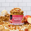 A jar of Sweet Earths All American Apple Pie Peanut Butter sits on a white surface with a white subway tile background. The jar is surrounded by ingredients that make up the product, including whole wheat crackers, white chocolate chips, dried apple pieces, and a sliced apple.