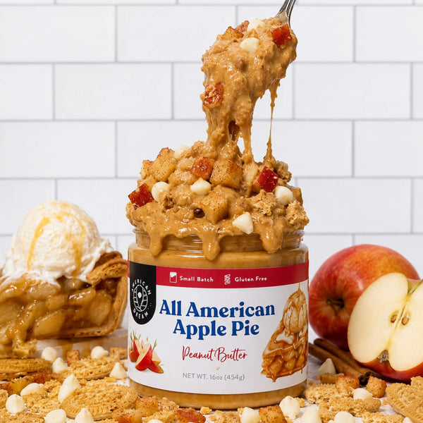 A jar of All American Apple Pie Peanut Butter sits on a white tiled surface with a spoon lifting a gooey, apple-pie flavored peanut butter spread. A scoop of vanilla ice cream appears on the left, and whole and sliced apples with cinnamon and cookie crumbs are arranged around the jar.