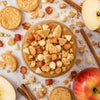 A glass bowl filled with a dessert or snack mixture of Oreo cookies, crumbled bread, white chocolate chips, and caramel-like sauce, surrounded by sliced apples, cinnamon sticks, and scattered Oreo cookies on a light surface.