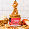 A jar of Sweetener Free All American Apple Pie Peanut Butter stands on a counter with a spoonful of the spread lifted high above the jar. A whole apple, a sliced apple, a scoop of vanilla ice cream, and cookie crumbs are arranged around the jar against a white subway tile wall background.