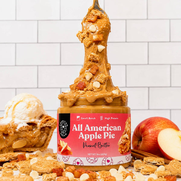A jar of Sweetener Free All American Apple Pie Peanut Butter stands on a counter with a spoonful of the spread lifted high above the jar. A whole apple, a sliced apple, a scoop of vanilla ice cream, and cookie crumbs are arranged around the jar against a white subway tile wall background.