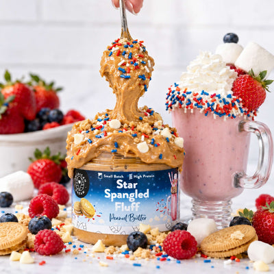 A jar of peanut butter topped with red, white, and blue sprinkles sits in front of a pink whipped drink with whipped cream, strawberries, blueberries, and cookie crumbs. A hand is visible holding a spoon above the jar, and the scene is styled with fresh berries, strawberries, raspberries, cookies, and sprinkles on a white surface.