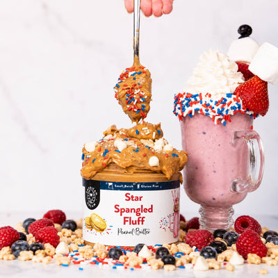A jar of Star Spangled Fluff Peanut Butter sits on a marble surface with whipped cream, strawberries, blueberries, popcorn, and red, white, and blue sprinkles around it. A spoon lifts peanut butter from the jar, and a pink milkshake with whipped cream and sprinkles stands beside it.